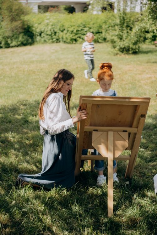 Free Teacher kneeling by child at easel, painting outdoors. Stock Photo