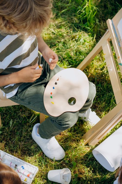 Free Young boy enjoys painting outdoors, holding a painter's palette on his lap, sunny day. Stock Photo