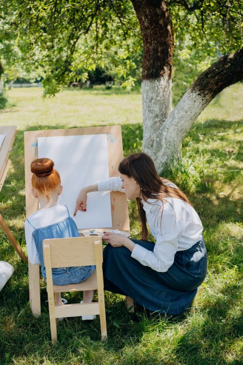 Free A teacher guides a young girl in an outdoor art class beneath a tree. Stock Photo