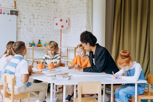 A Woman and a group of Children Sitting at a Table with Books and Pens