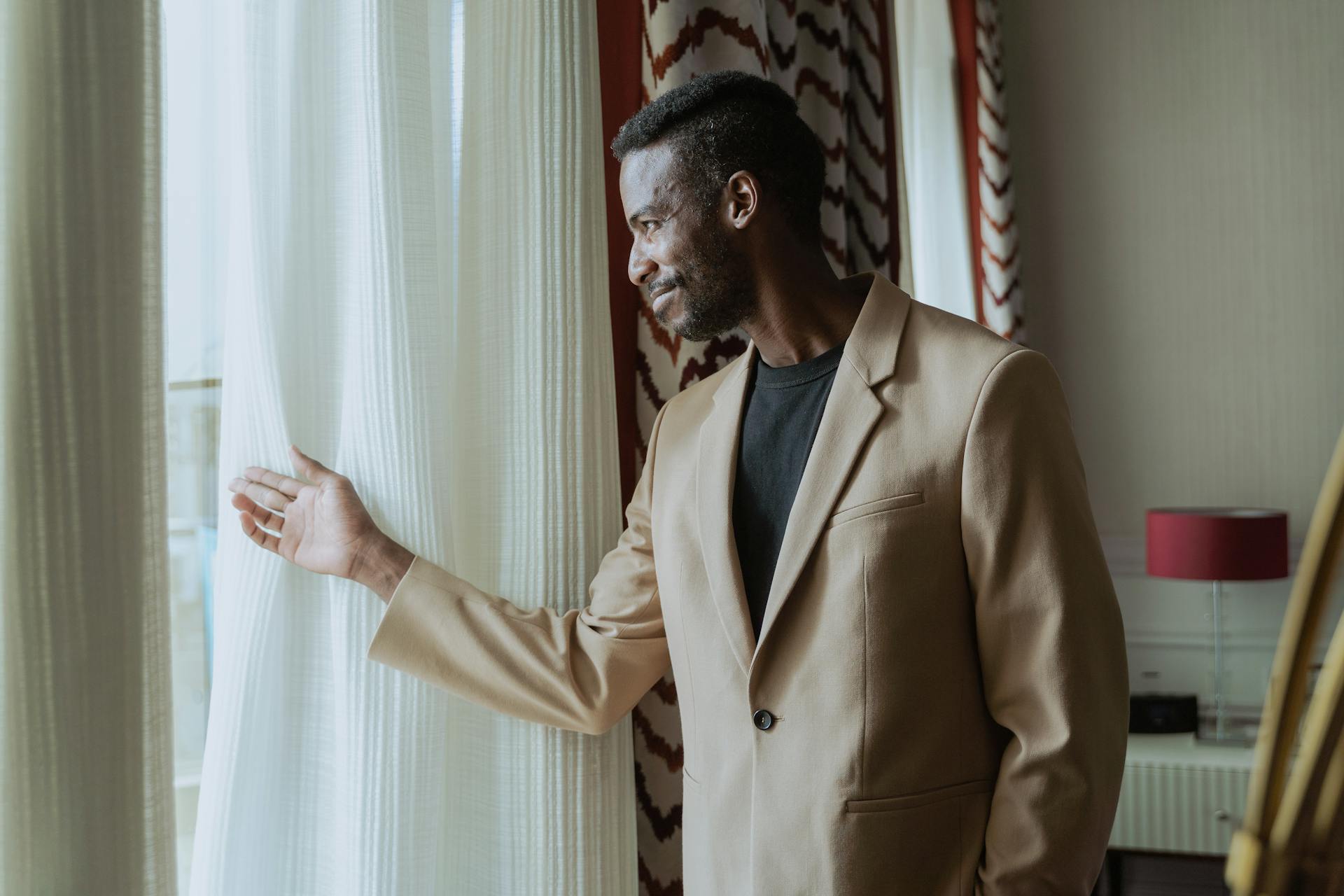 African American man in tan suit looking outside through window in a cozy indoors setting.