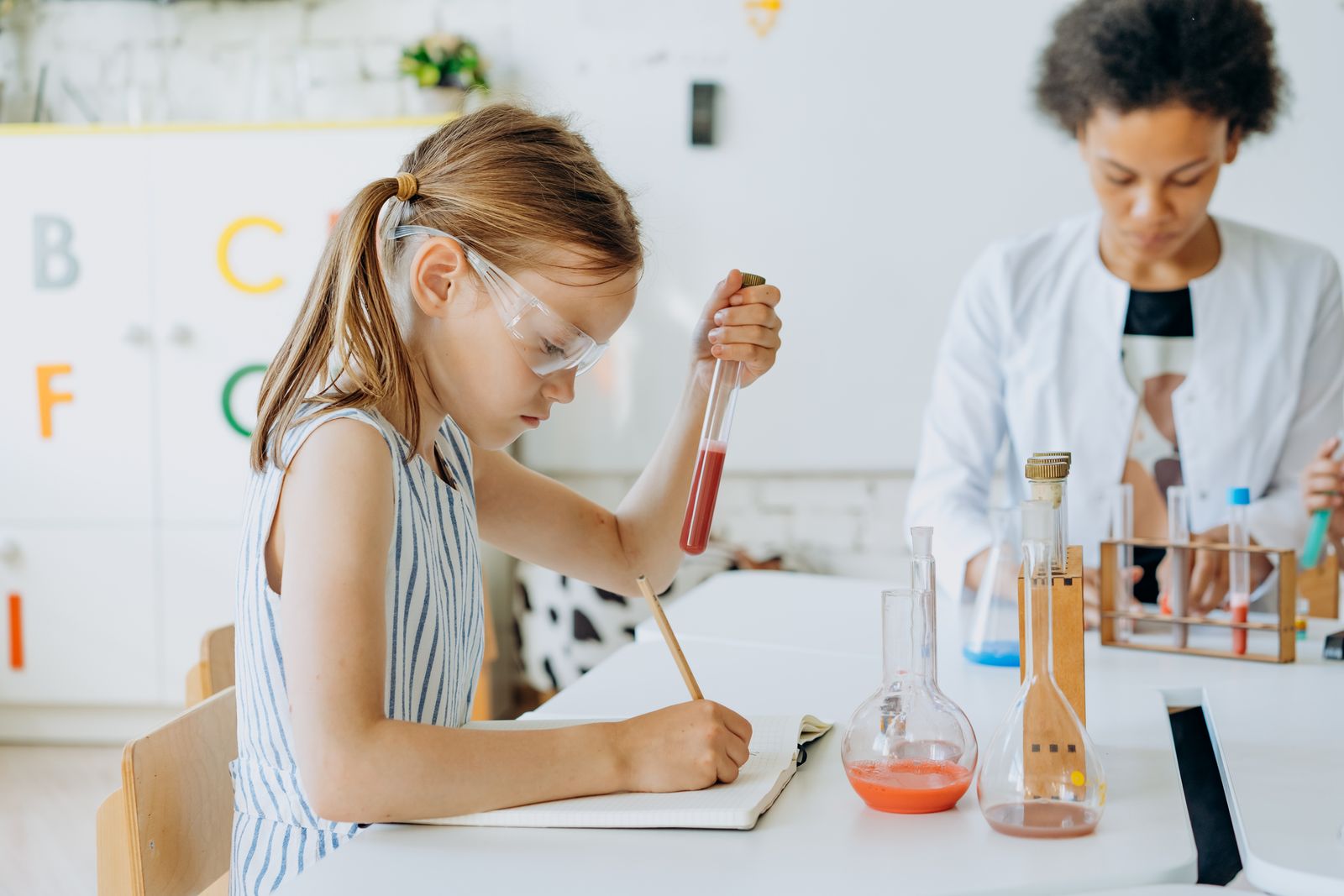 A child holding a test tube and writing observations during a supervised classroom science experiment