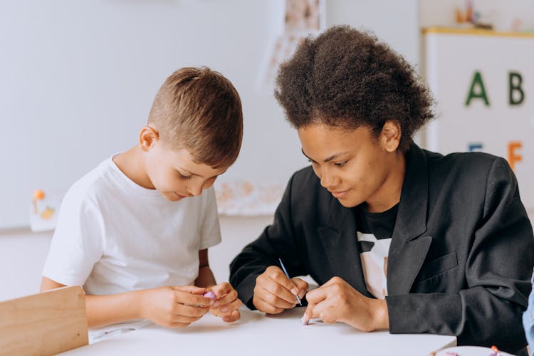 A Boy In White Shirt Sitting At The Table With His Teacher