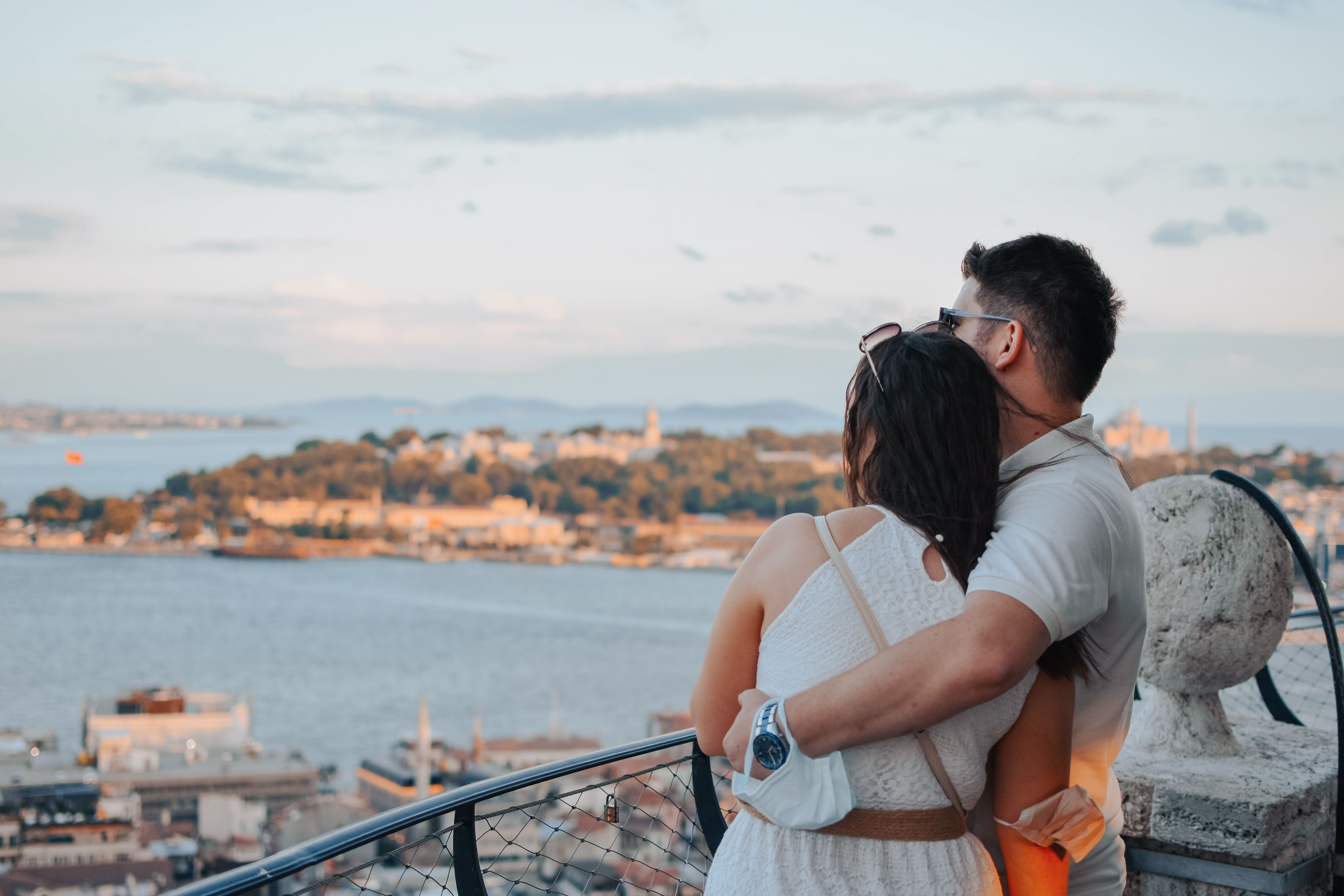 Casal abraçando em varanda com vista para cidade costeira europeia, luxo e romance.