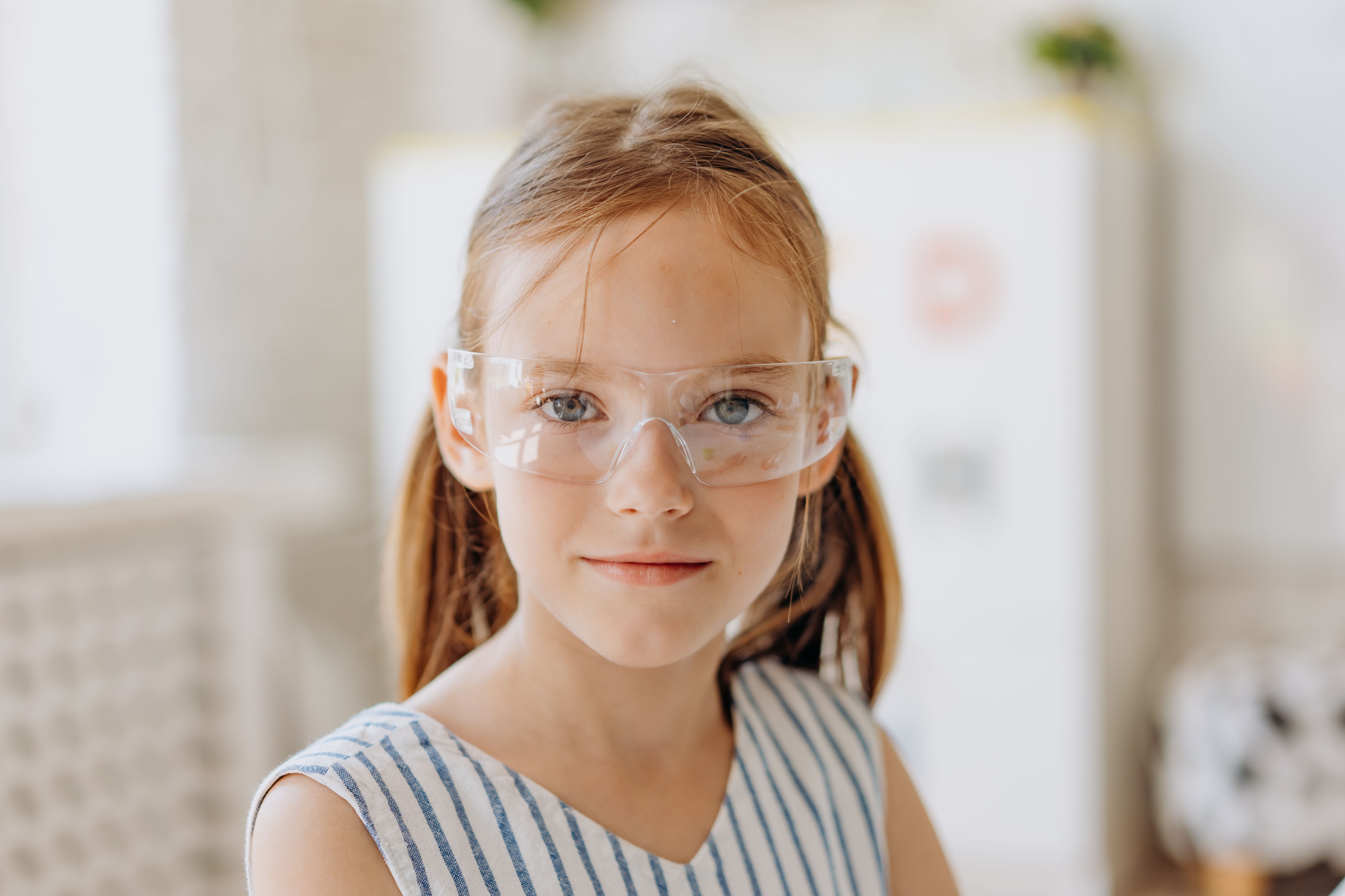 A young girl in a striped dress and safety glasses standing in a bright classroom.