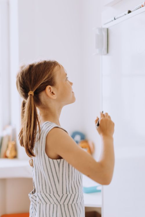 Free Side view of a young girl with pigtails writing on a whiteboard in a classroom setting. Stock Photo