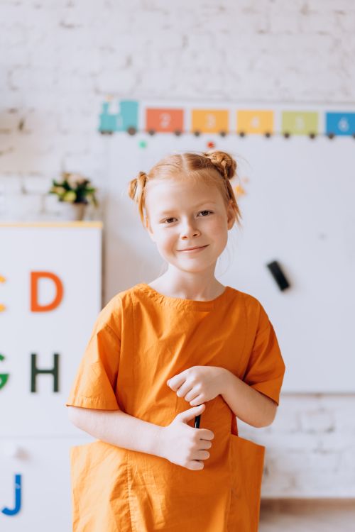 Free A young girl wearing an orange dress smiles at the camera in a bright room. Stock Photo