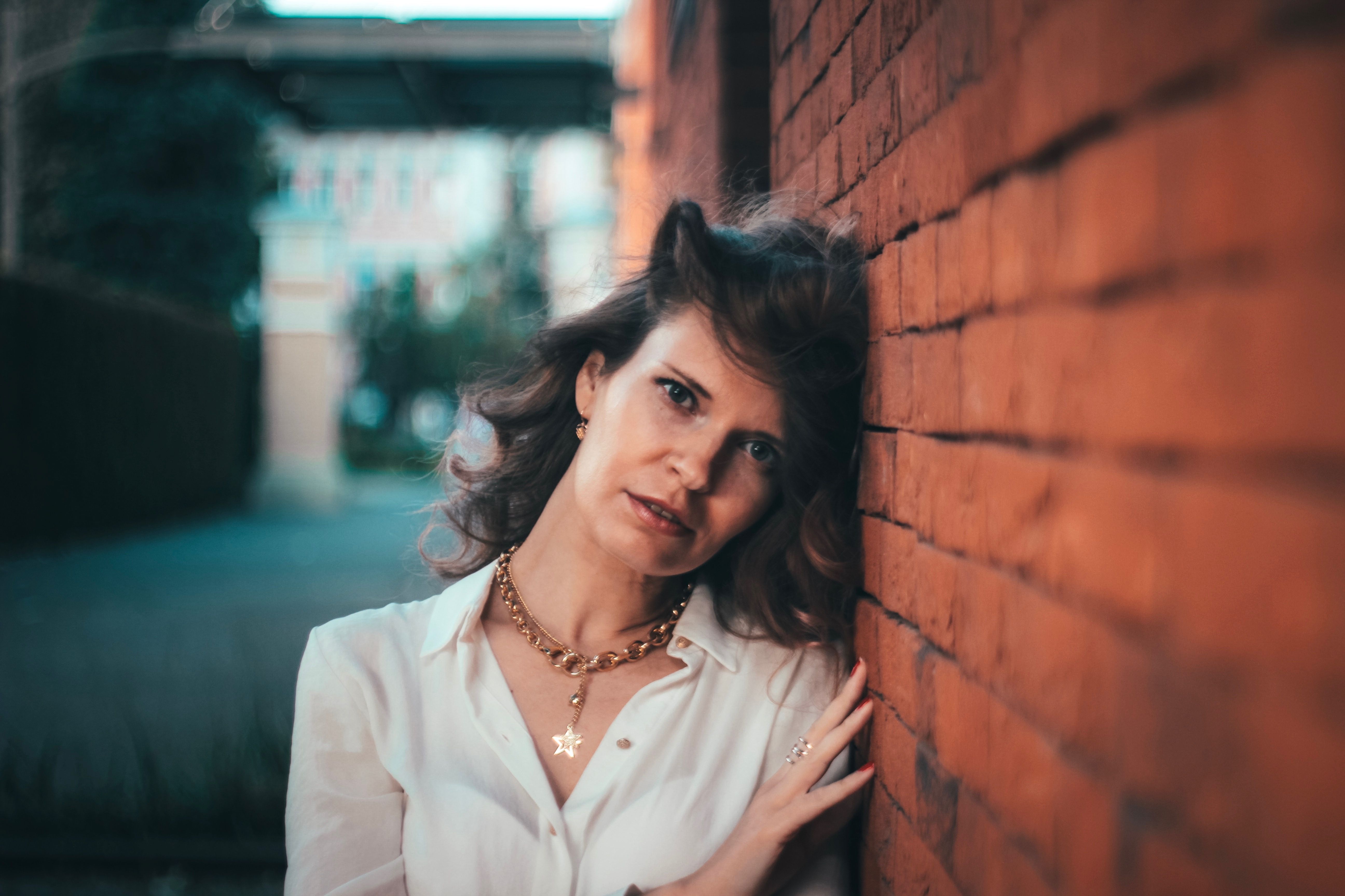 Elegant woman in a gold necklace and white shirt leaning against a brick wall. Blurred urban background.