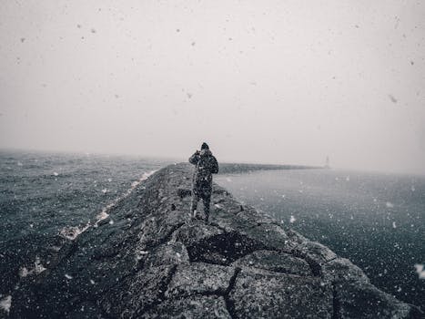 A person walks along a breakwater during a snowy day by the sea, creating a moody atmosphere.