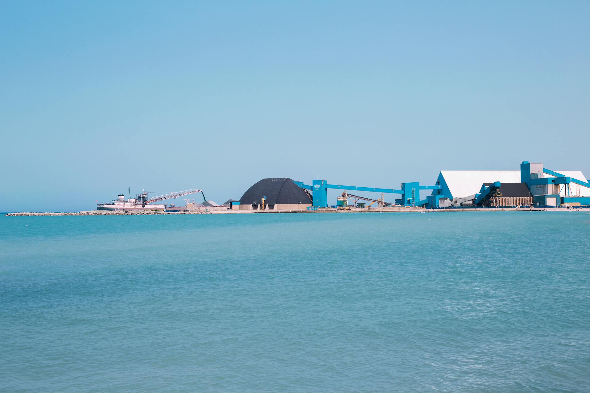 A tranquil view of an industrial port surrounded by calm blue waters, featuring a cargo ship and cranes in the distance.