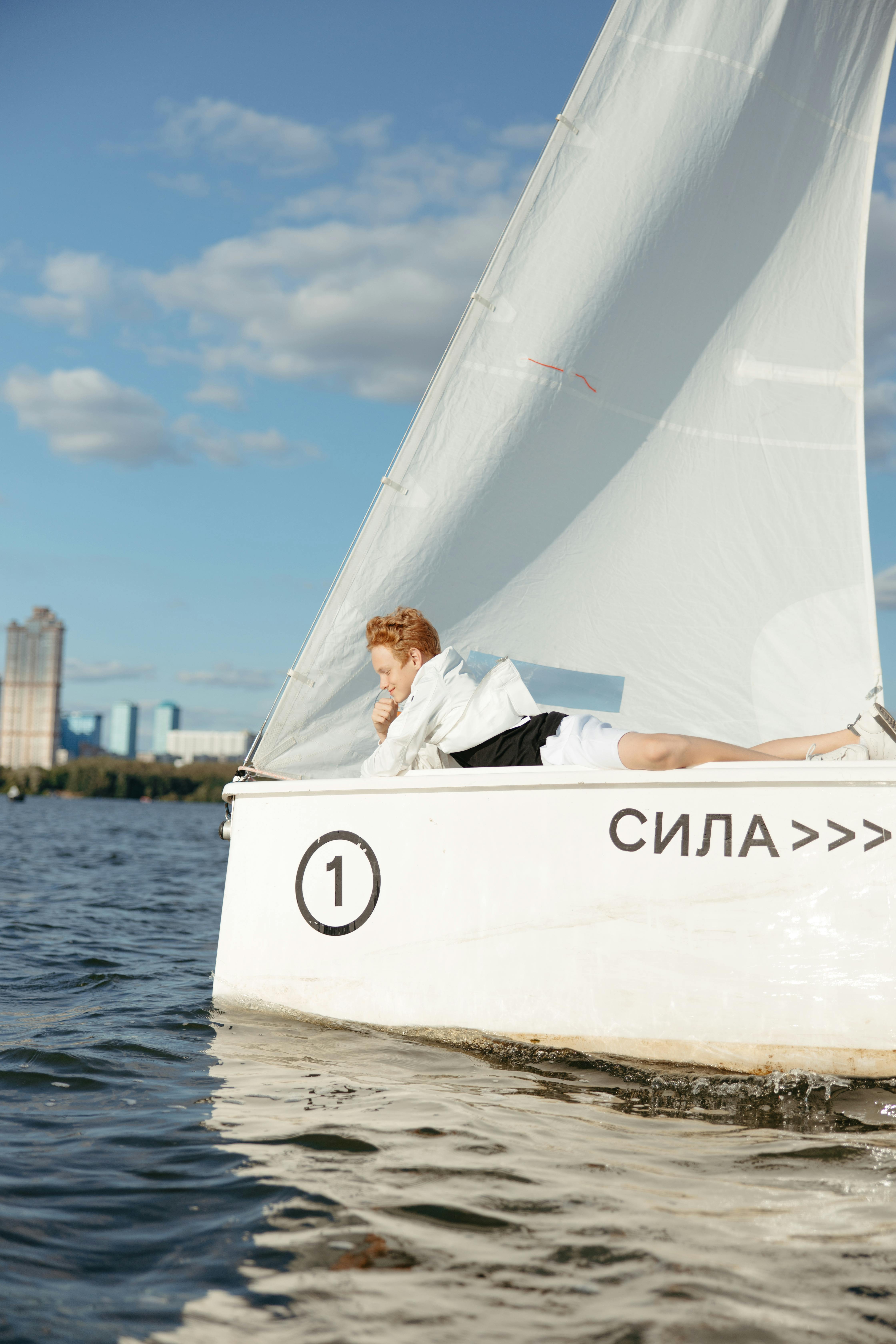 A person lounging on a sailboat with clear skies and cityscape in the background.