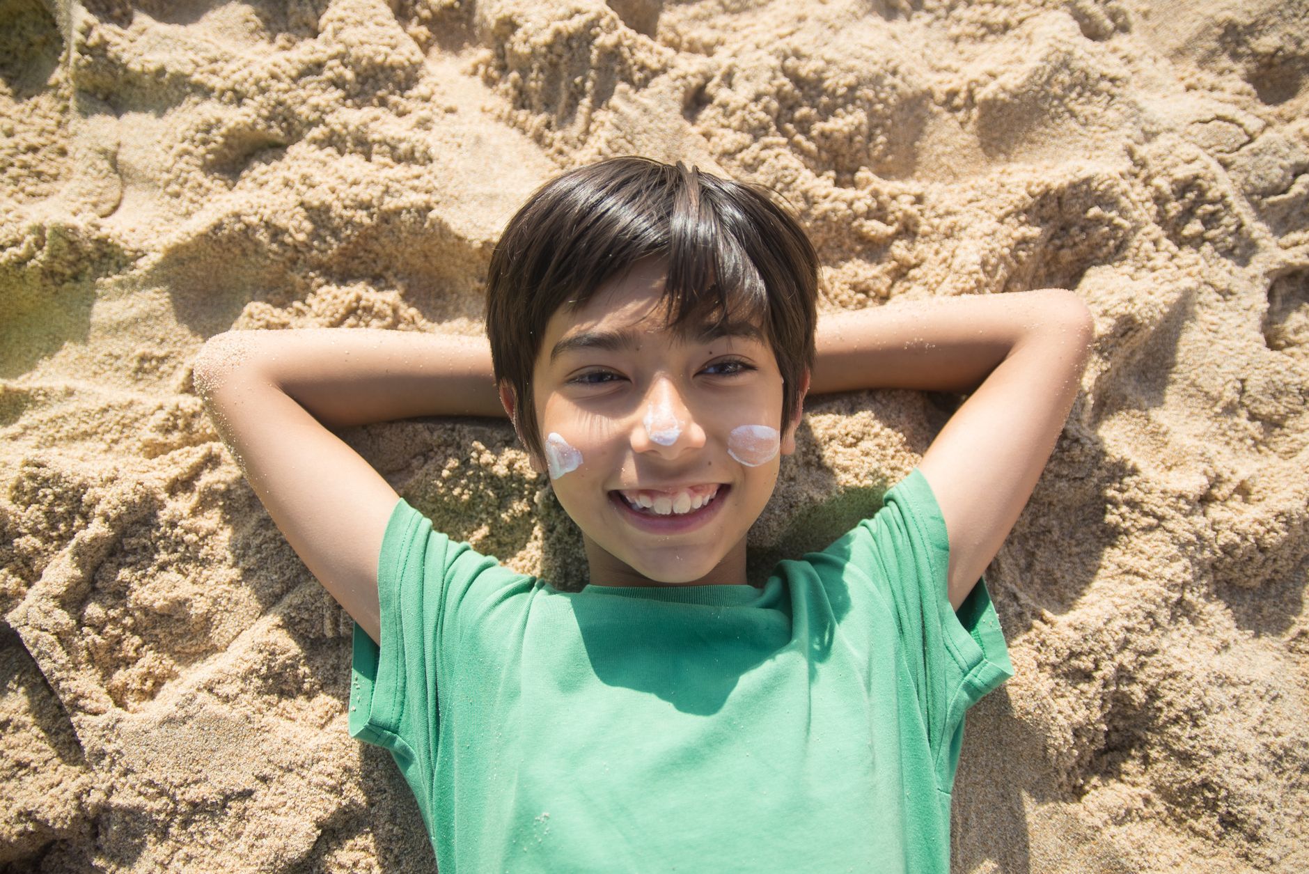 A cheerful boy lies on the sand at the beach, smiling with sunscreen on his cheeks. Perfect summer day.