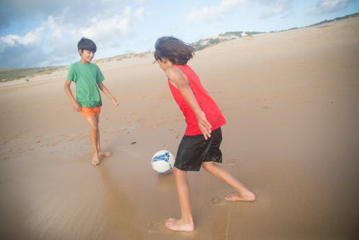 Two children playing soccer barefoot on a sunny Portuguese beach during summer, enjoying outdoor fun.
