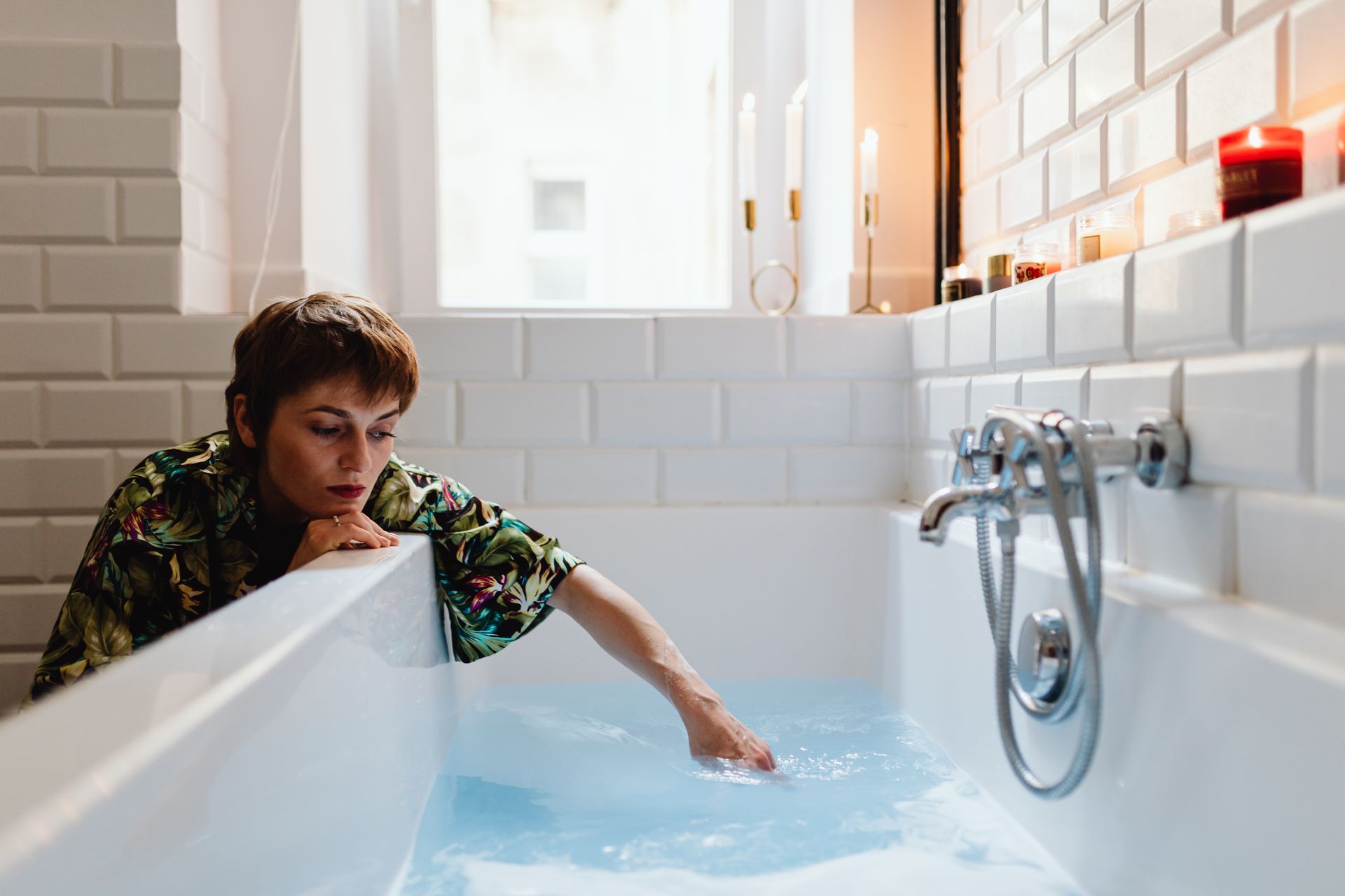A woman in a serene bathroom touches the water in a modern white tiled bathtub.