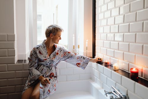 A Woman in Floral Bathrobe Sitting on a Bathtub while Holding a Lighted Candle