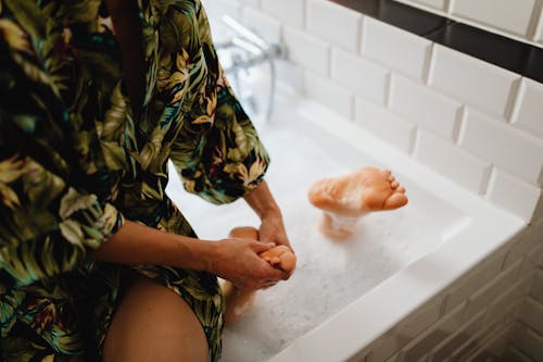 Person Massaging a Feet While Sitting on a Bathtub Wearing a Printed Robe