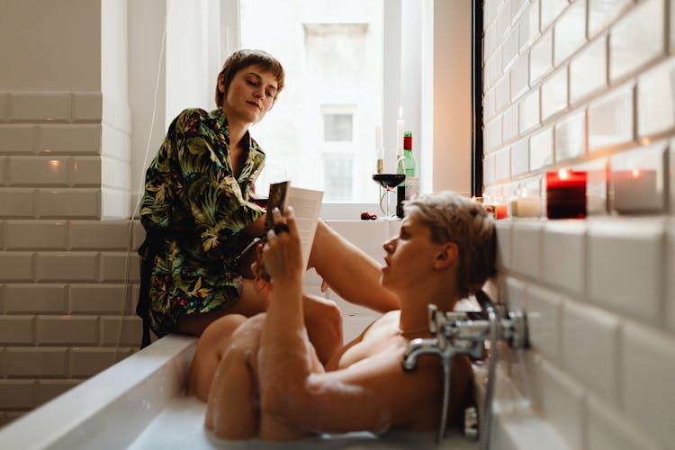 Woman Sitting On The Edge Of The Bathtub Next To Her Girlfriend Reading In The Bath