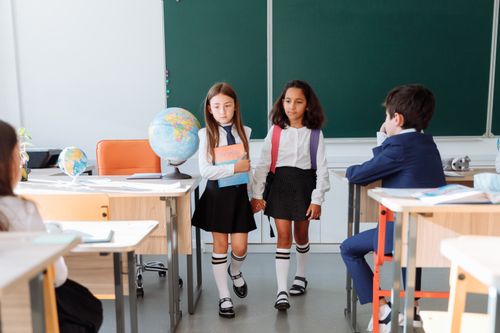 Free Two girls walking hand in hand inside a classroom with students and a globe. Stock Photo