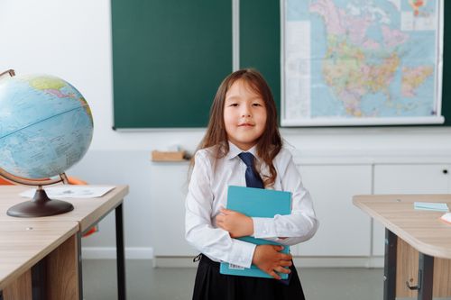 Free Smiling young girl in school uniform holding a book in a classroom setting. Stock Photo