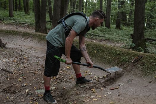 A man shoveling dirt on a forest trail, creating a path surrounded by greenery.