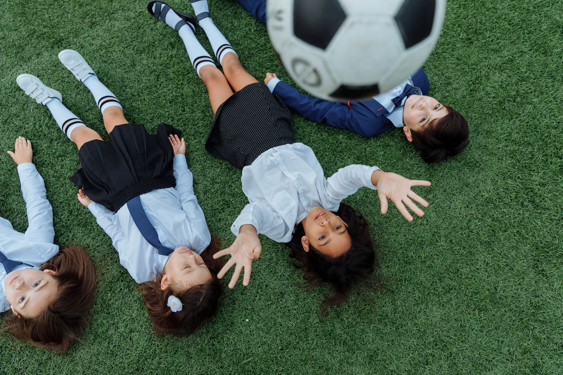 A group of young children lying on grass reaching for a soccer ball during play