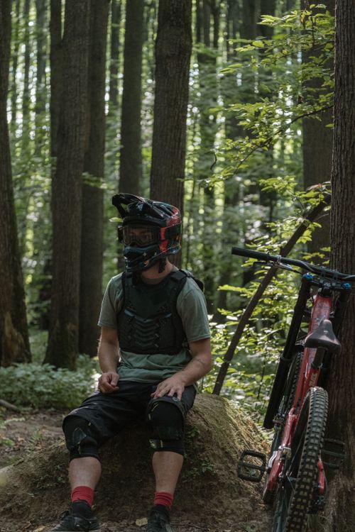 Free A mountain biker wearing a helmet takes a break in a dense forest with a bicycle nearby. Stock Photo