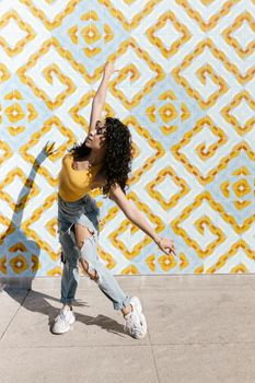 Woman in a dynamic dance pose against a patterned wall in sunlight, showcasing style and freedom.