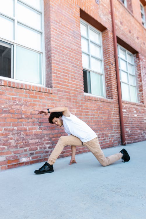Photo of a Man in a White Shirt Dancing Beside a Brick Wall