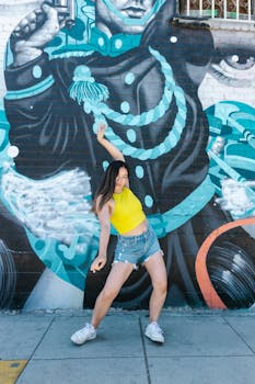 Young woman in a yellow tank top dances energetically against a colorful graffiti wall.