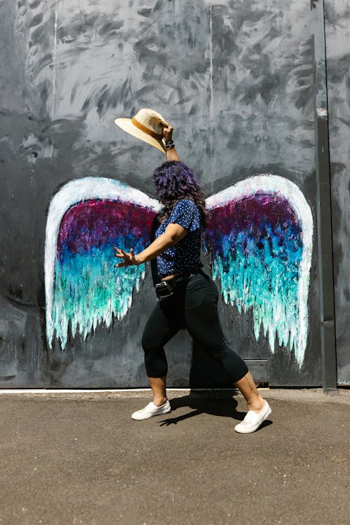 Free A Woman Holding a Hat Posing by a Wall with Angel Wings Stock Photo