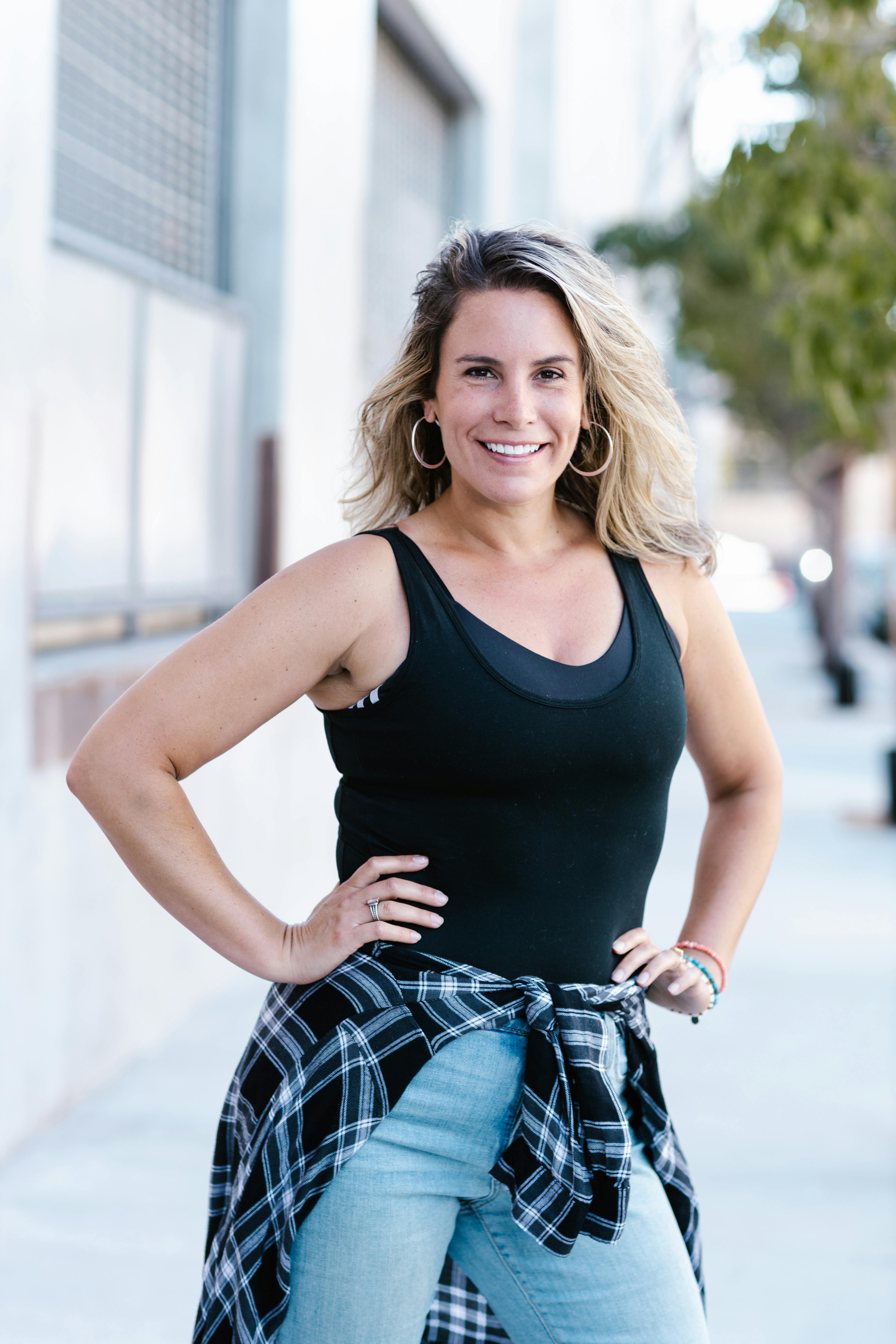 Smiling woman with hands on waist, wearing tank top and jeans, outdoors.