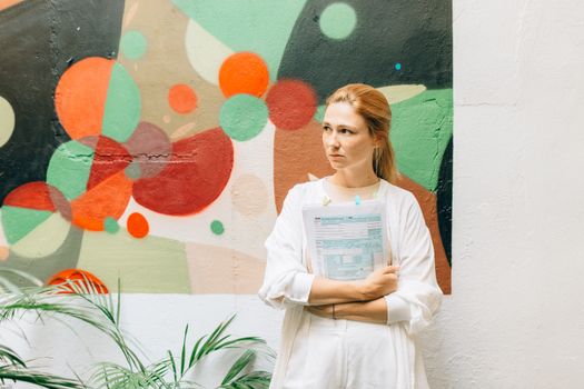 A woman in a white outfit standing indoors holding documents, with a colorful abstract mural in the background.