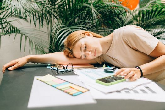 Photo by Nataliya Vaitkevich A tired woman calculating her taxes at a desk covered with papers and a smartphone.