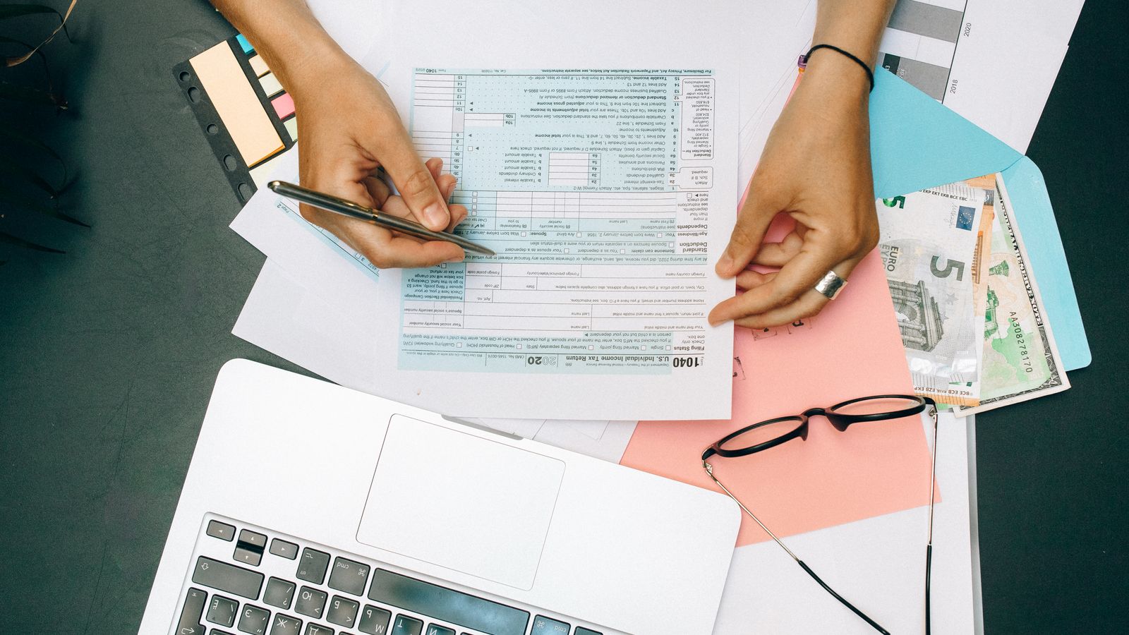 Hands writing on tax documents with laptop, glasses, and currency on desk.