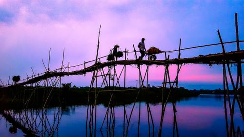 Free A person with a bicycle crosses a bamboo bridge reflecting on the calm river during twilight. Stock Photo