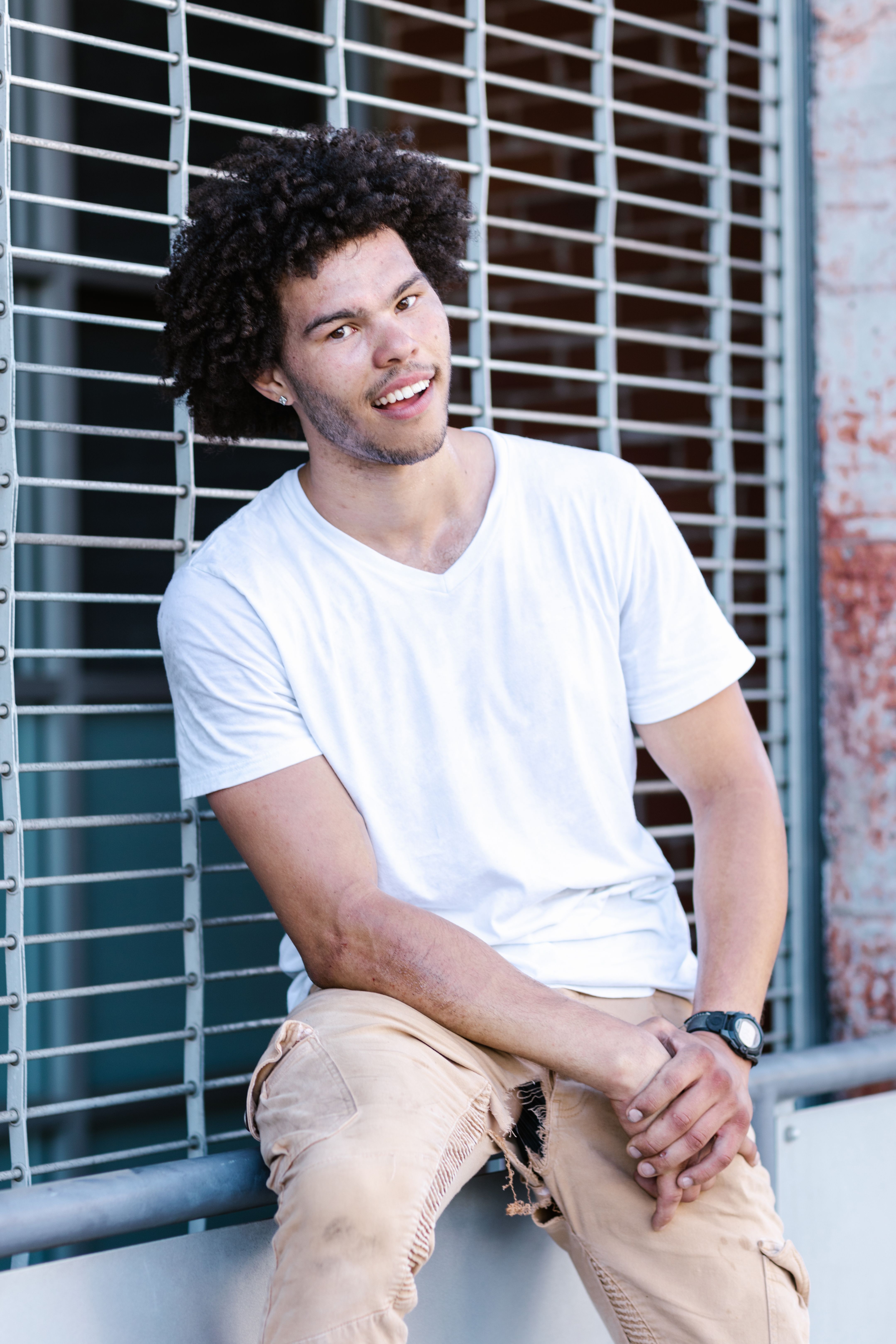 Young man with curly hair sitting casually against a metal railing outdoors.