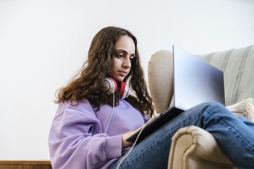 Young woman wearing headphones, sitting on a couch and using a laptop, indoors.
