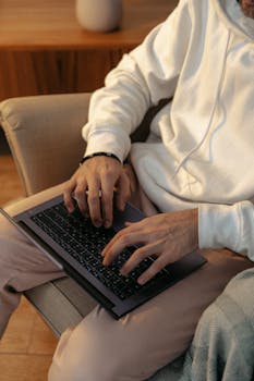 Close-up of a man typing on a laptop while sitting on an armchair in a cozy setting.