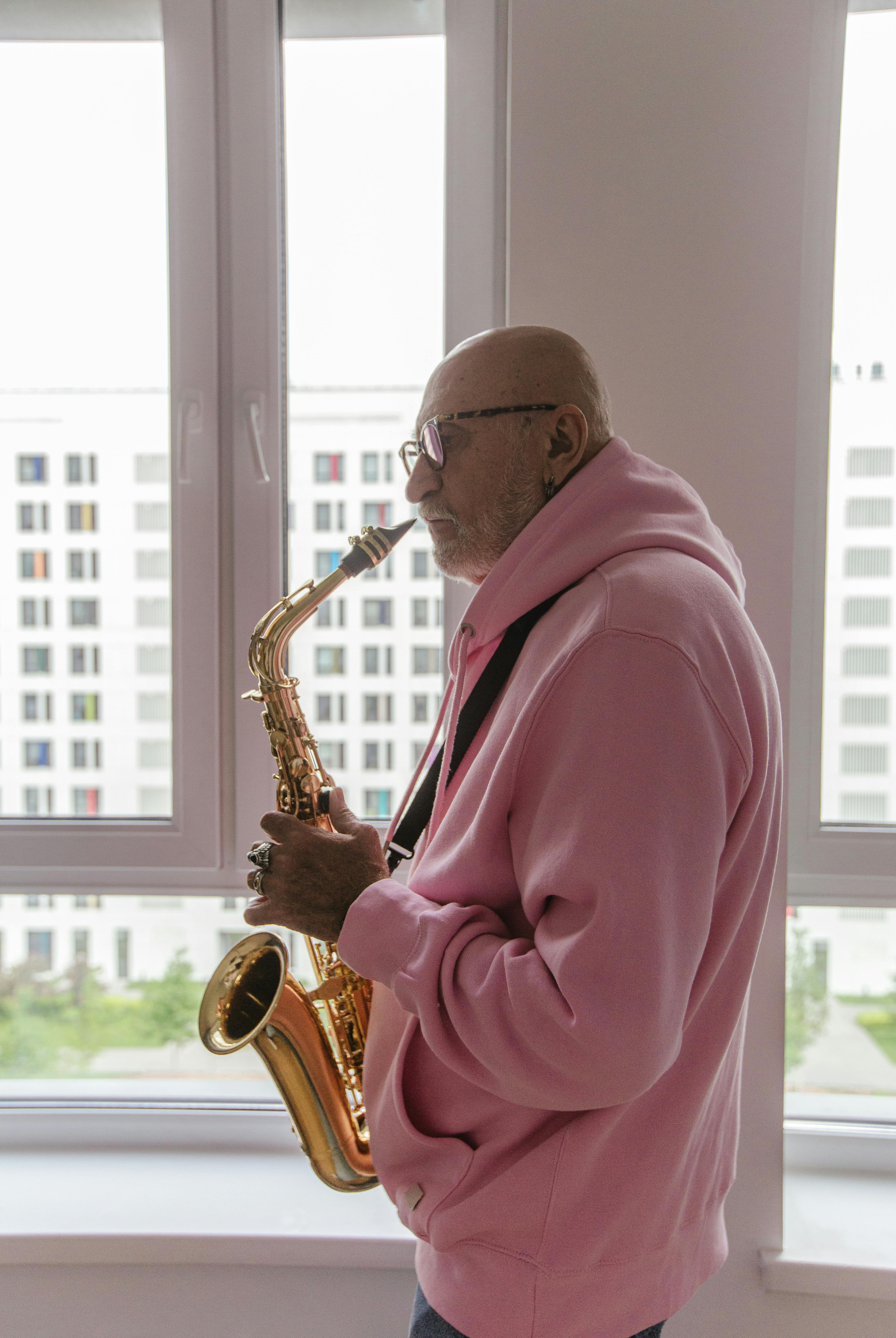 Elderly musician playing saxophone indoors near a bright window. Side view.