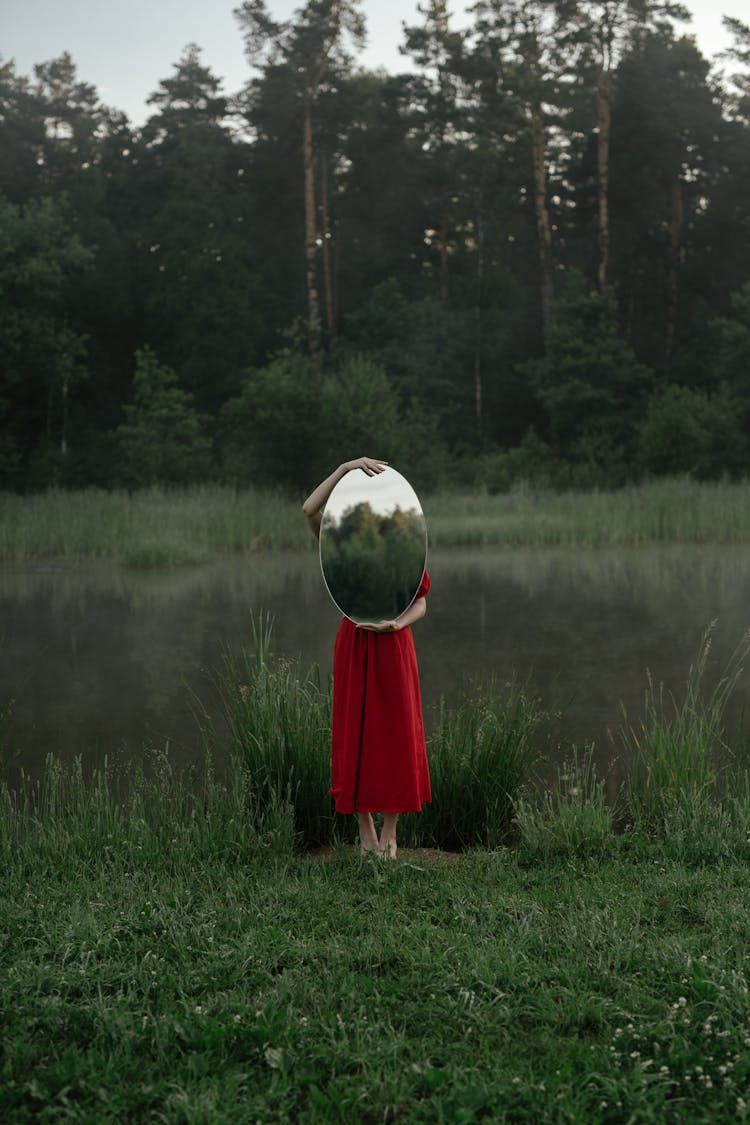 A Person In A Red Dress Holding A Mirror Near A Body Of Water