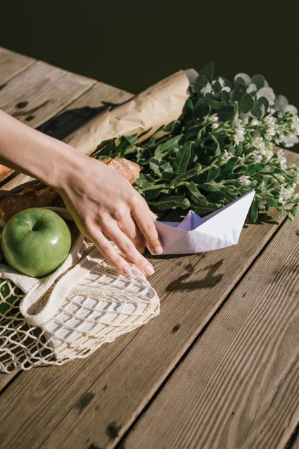 A serene arrangement with a hand reaching for a paper boat among a bouquet, apple, and groceries on a rustic table.