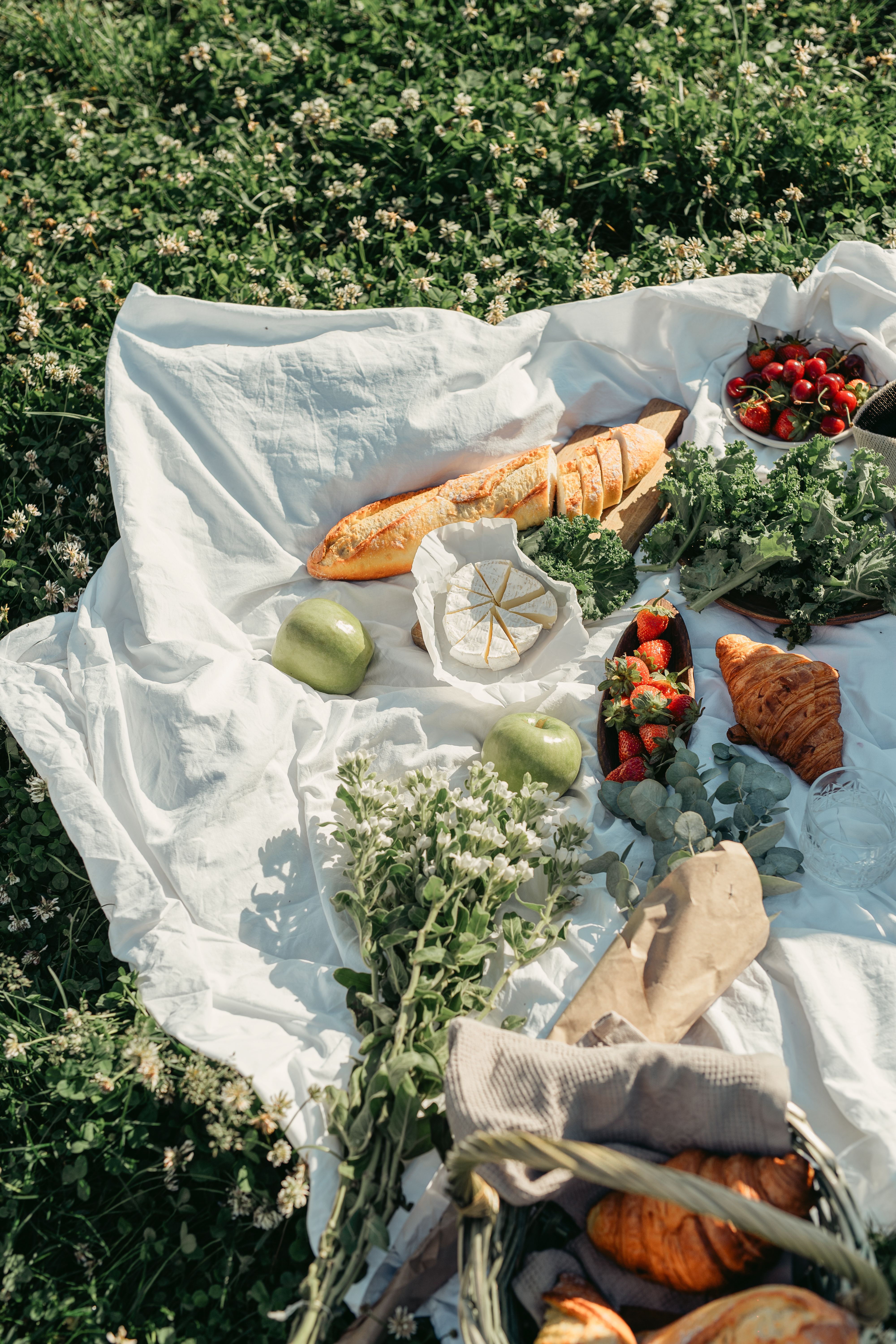 Charming picnic setup with fresh baguettes, croissants, fruits, and flowers on a sunny day.