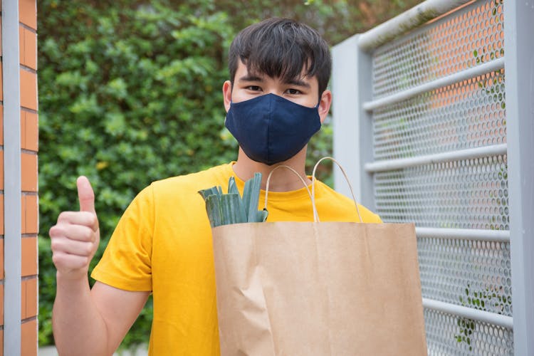 A Man In A Face Mask Holding A Paper Bag Of Groceries