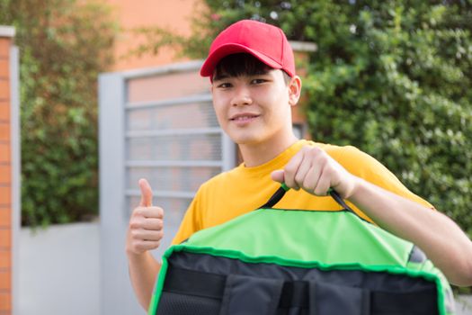 Asian delivery man in red cap and yellow shirt giving thumbs up while holding a bag outdoors.