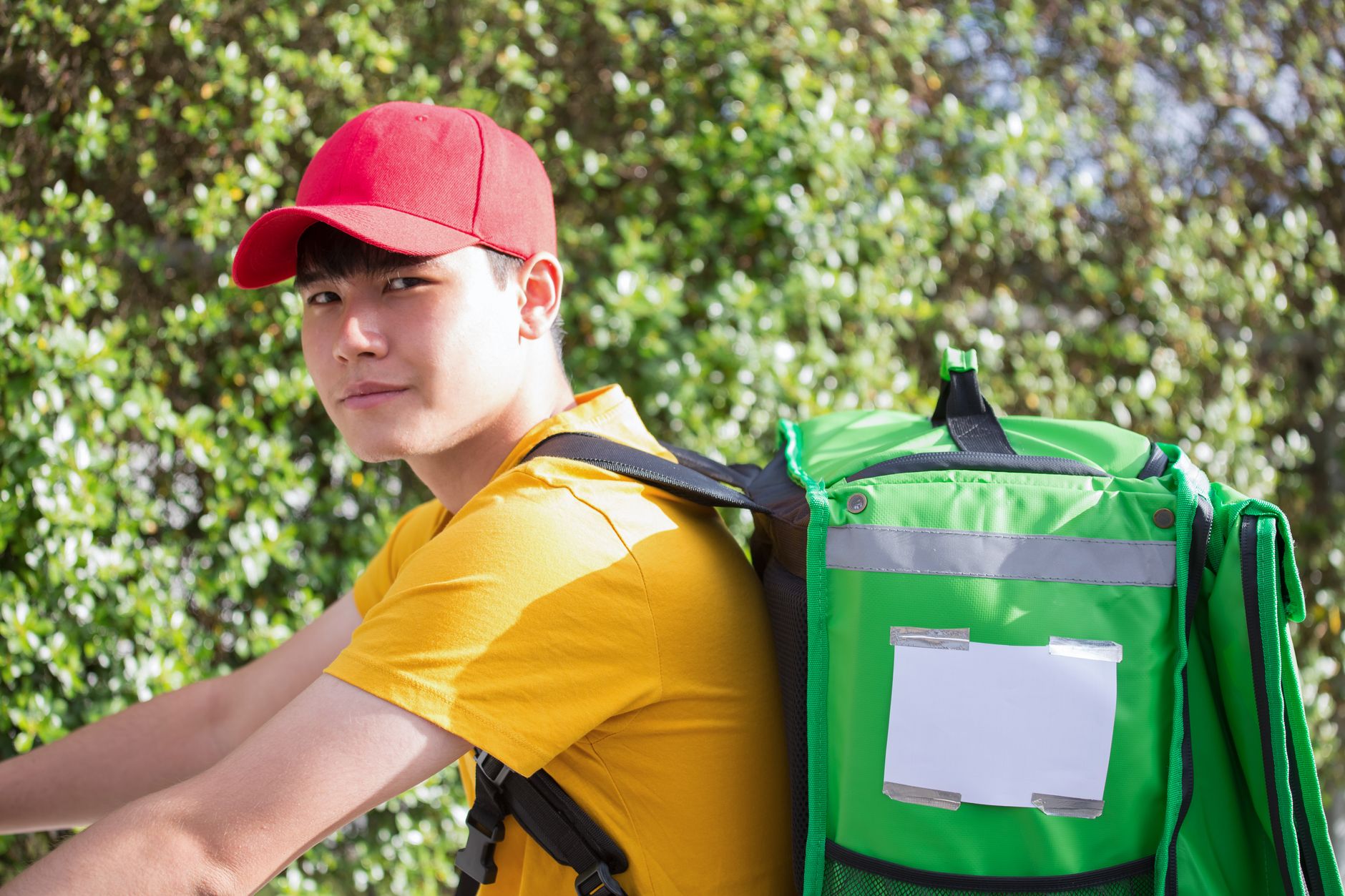 Asian delivery man wearing a red cap with an insulated bag, ready for delivery outdoors.