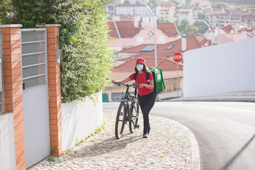 A female bicycle courier with an insulated bag walks on a cobblestone street in Portugal, wearing a mask.