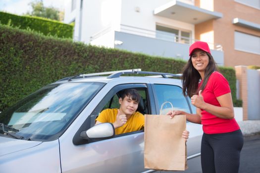 Asian delivery woman handing paper bag through car window with man inside, suburban Portugal setting.