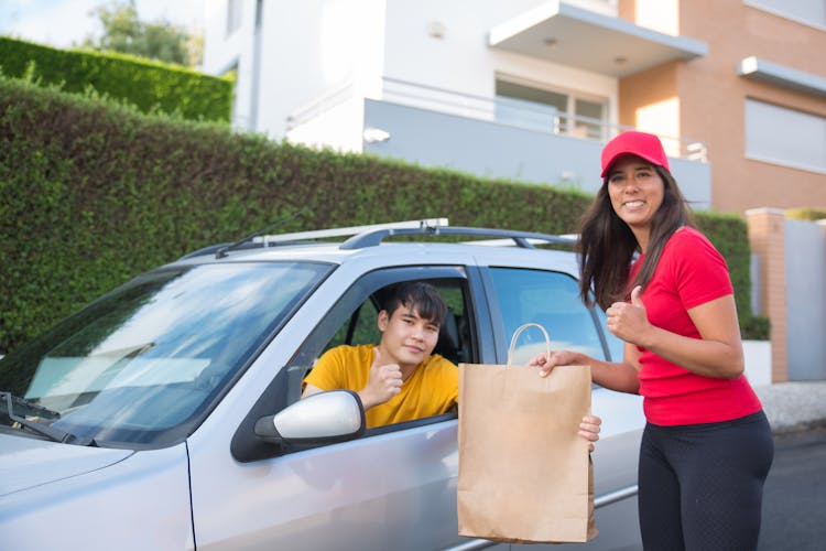 Woman In Red Shirt Handing Brown Paper Bag To The Man Sitting Inside A Car