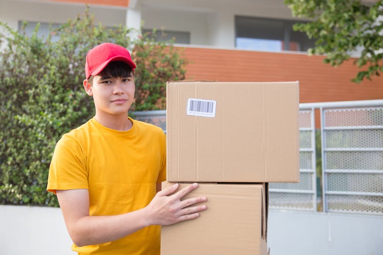 A Man In Yellow Shirt And Red Cap Carrying Stacks Of Cardboard Boxes While Looking At The Camera