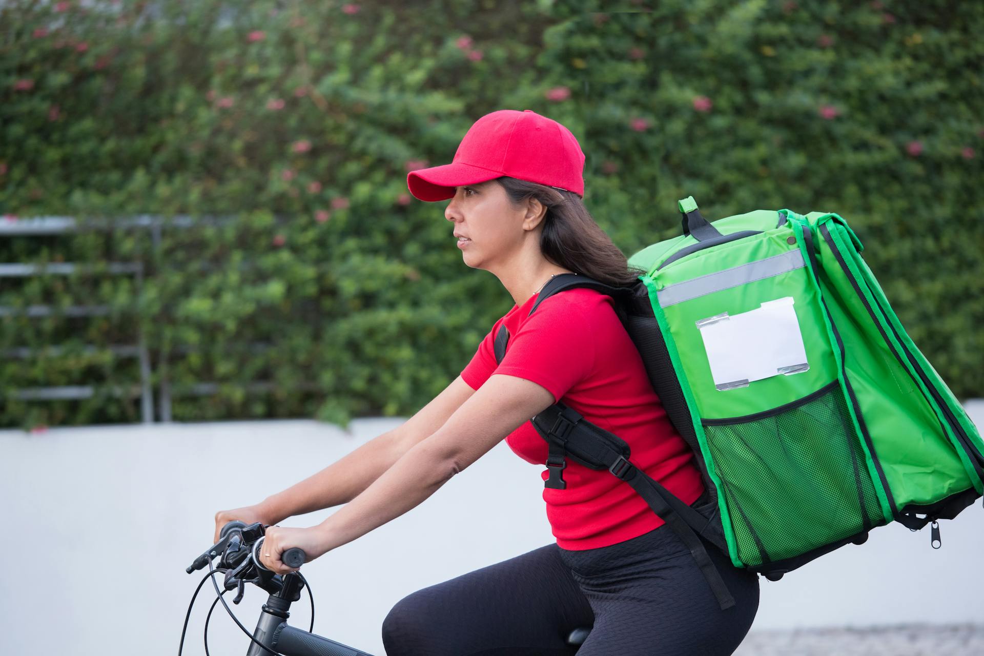A female courier wearing a red cap rides a bicycle with a large green delivery bag outdoors.